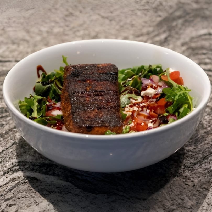 A white bowl filled with a dark seared salmon fillet on a bed of greens, tomatoes, and feta.