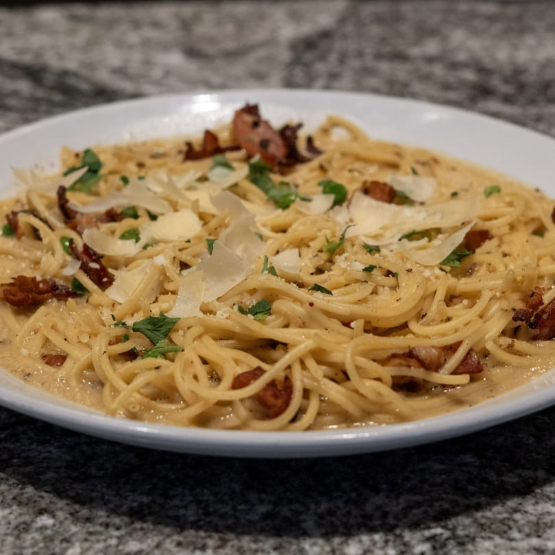 Creamy spaghetti carbonara with crispy bacon, fresh parsley, and shaved Parmesan cheese on a white plate.