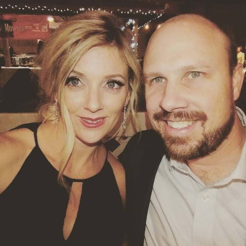 A smiling blonde woman and a bearded man pose for a selfie in a dimly lit restaurant.