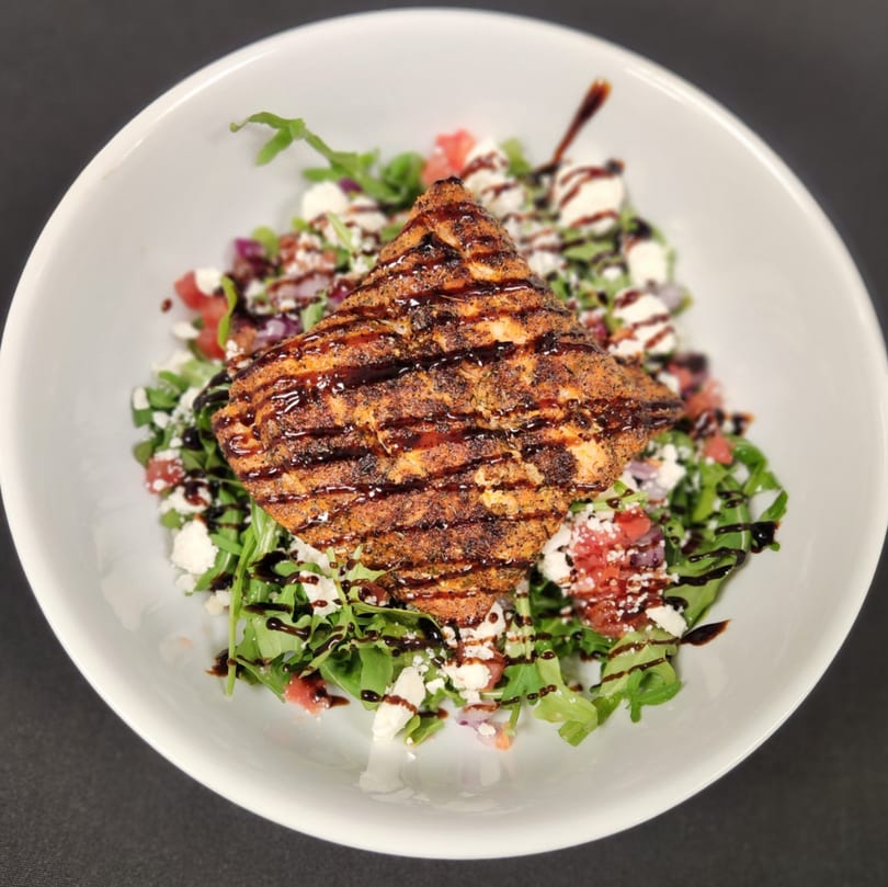 Grilled salmon with balsamic glaze on a bed of arugula, feta, and tomatoes in a white bowl.