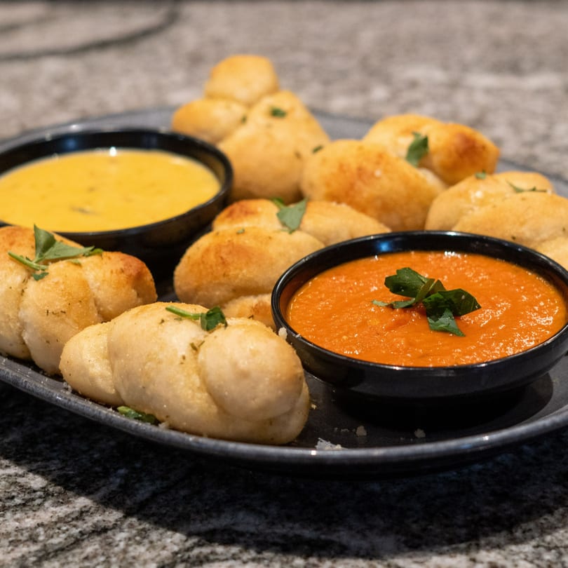 Garlic knots with two dipping sauces, one yellow and one red, on a dark plate.