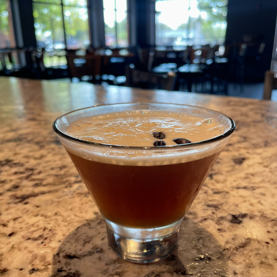 Espresso martini with coffee beans on top, on a speckled bar counter with a restaurant in the background.